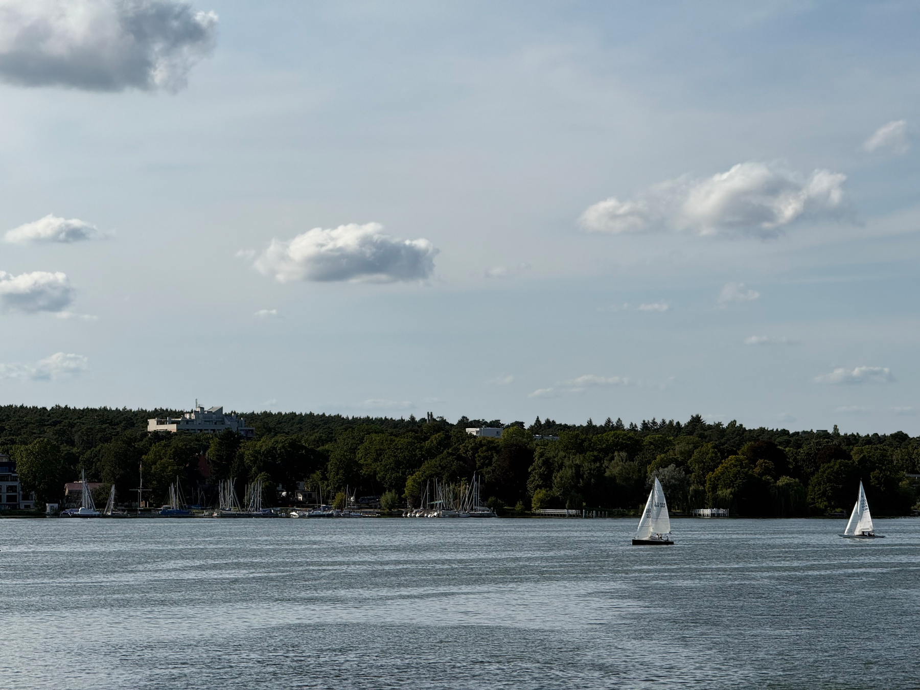 Auf einem ruhigen See segeln zwei kleine Segelboote, während im Hintergrund ein bewaldetes Ufer mit Gebäuden zu sehen ist.