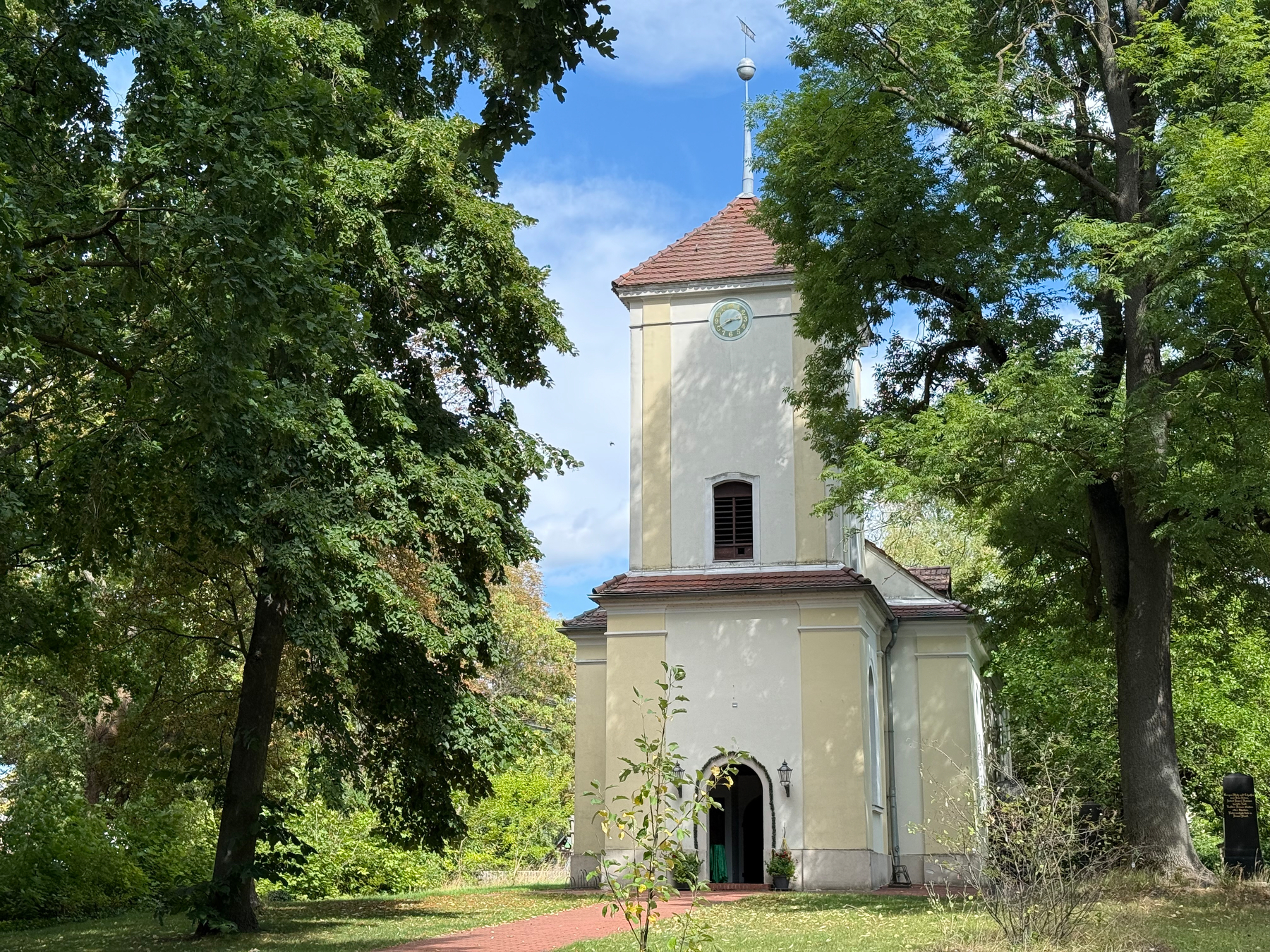 Eine kleine hellgelbe Dorfkirche mit einem Turm und einem Zifferblatt, umgeben von hohen Bäumen, steht unter einem blauen Himmel.