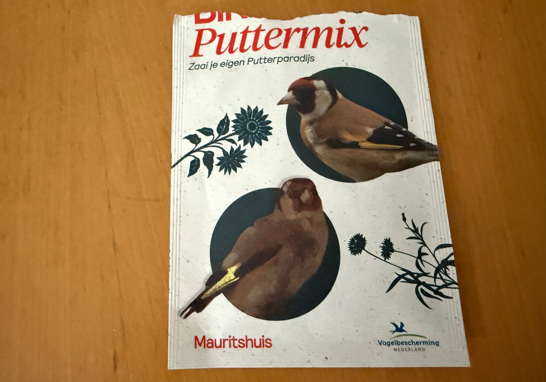 An opened paper sachet with a drawing of 2 finches that says Puttermix (Dutch text “Zaai je eigen putterparadijs”) on a wooden tabletop